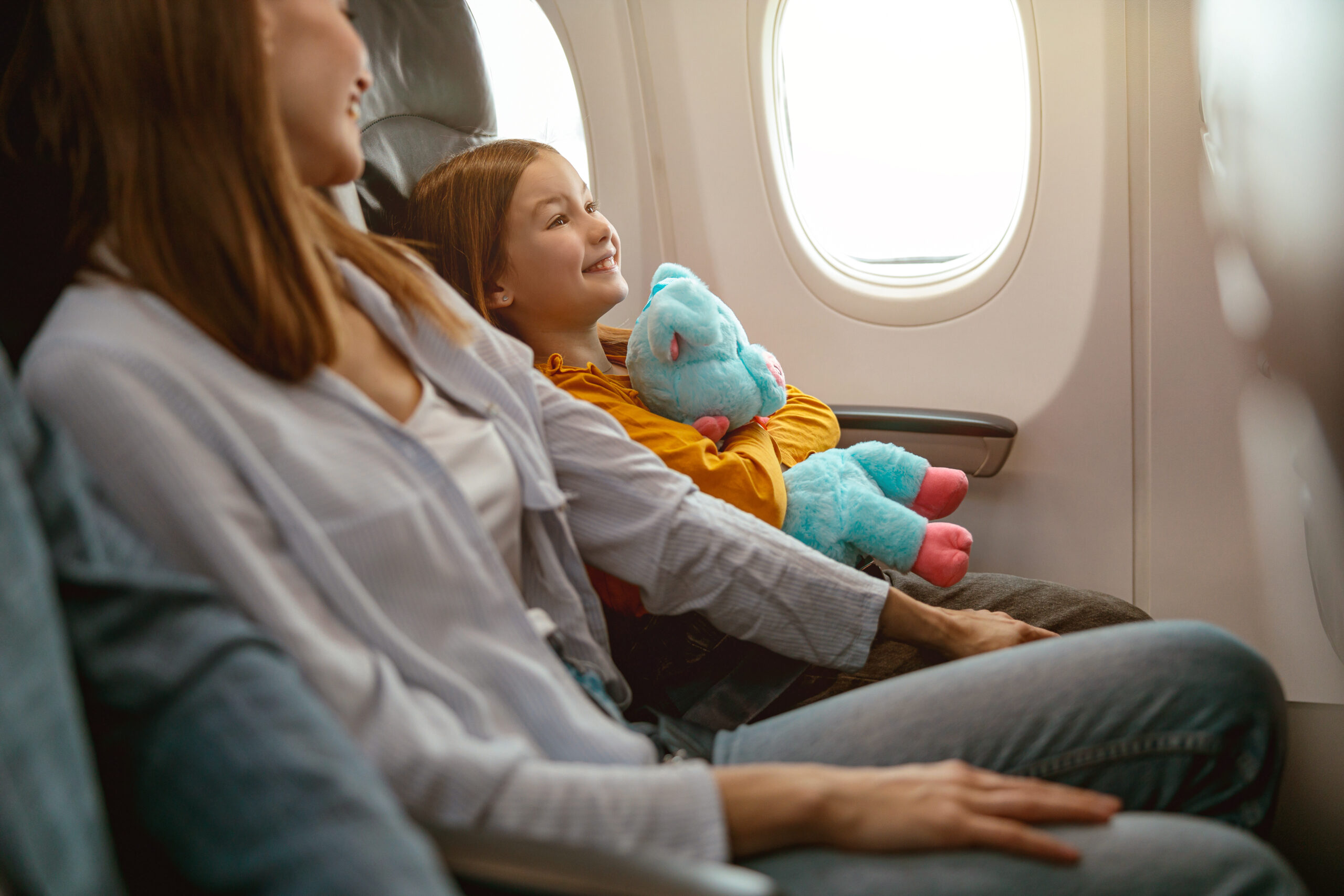 Cheerful little girl holding toy and smiling while sitting next to woman during flight in aircraft