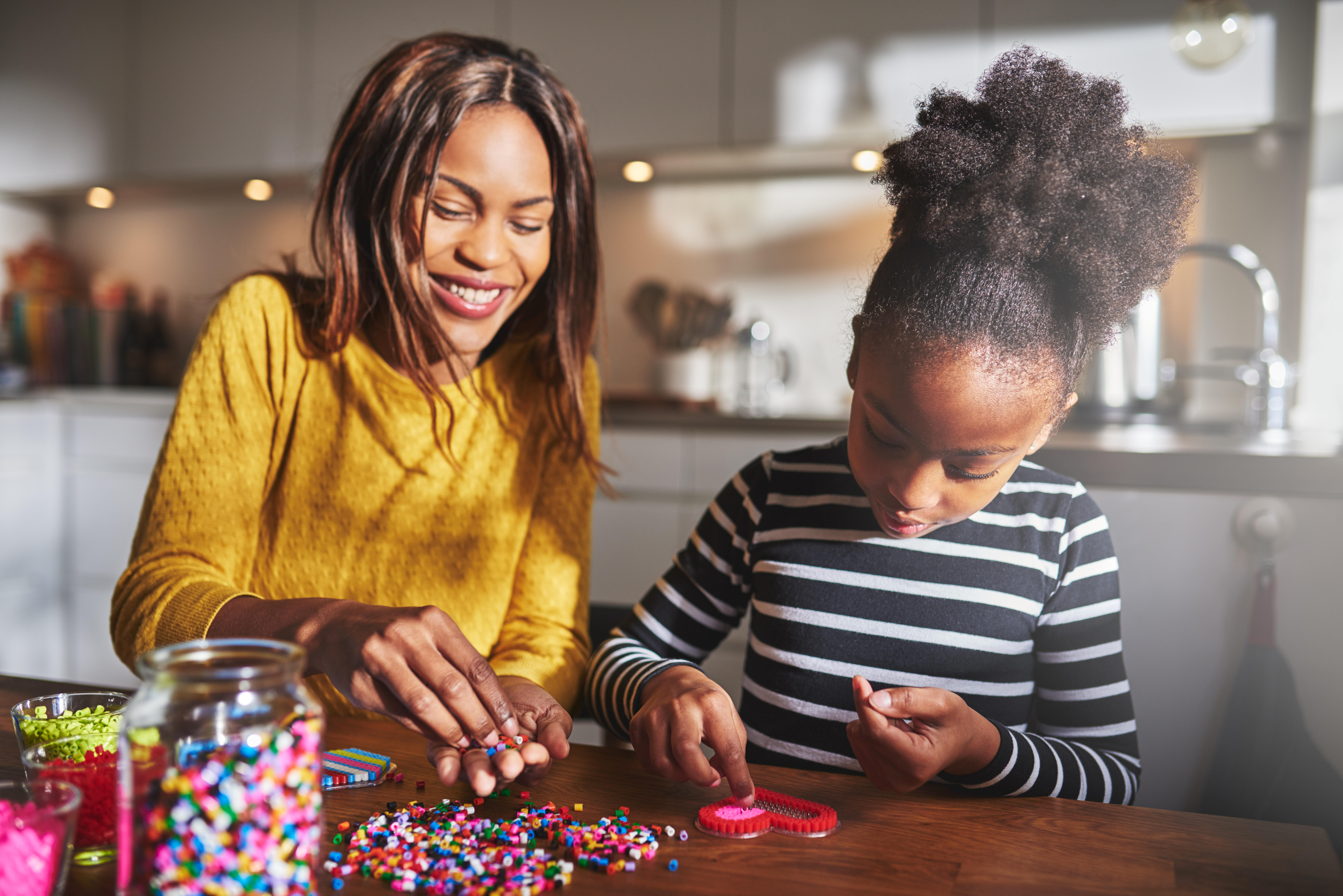 Female parent helping cute child in striped shirt choose bead colors for heart shape frame on table in kitchen